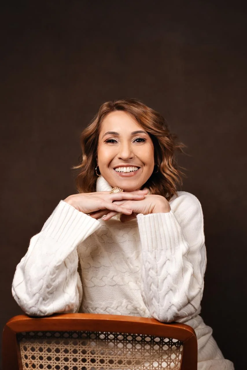 Lorenna Torres sorrindo, de suéter claro, com as mãos apoiadas no encosto de uma cadeira